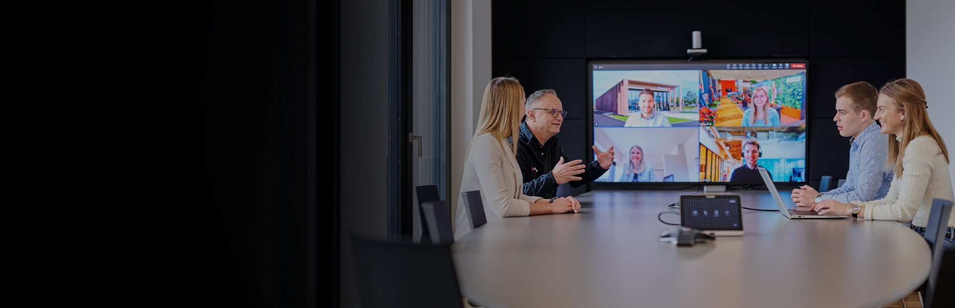 Five people in a meeting room in front of a screen showing an active video conference.