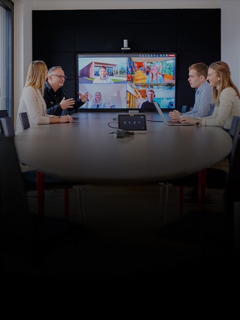 Five people in a meeting room in front of a screen showing an active video conference.