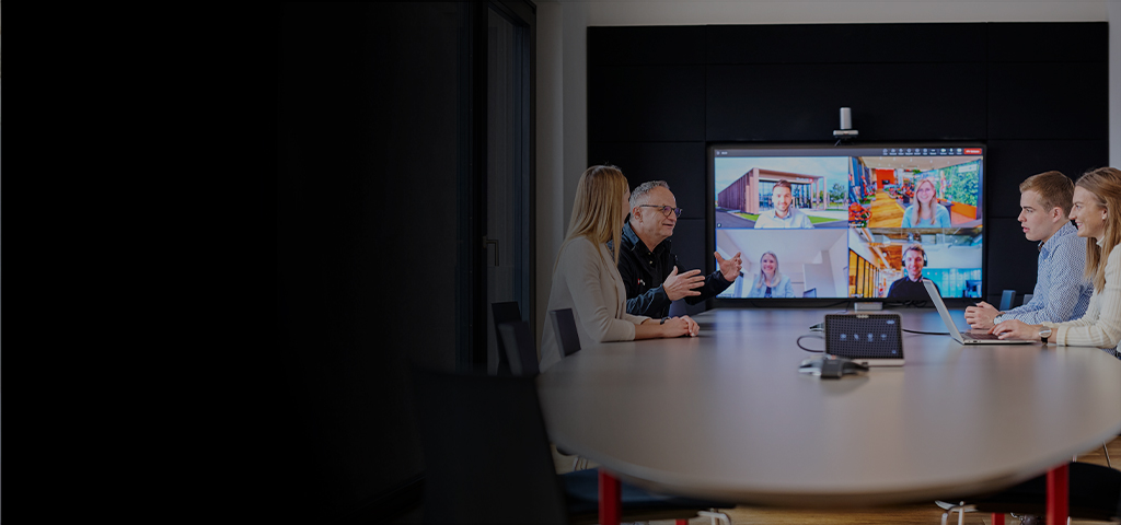 Five people in a meeting room in front of a screen showing an active video conference.