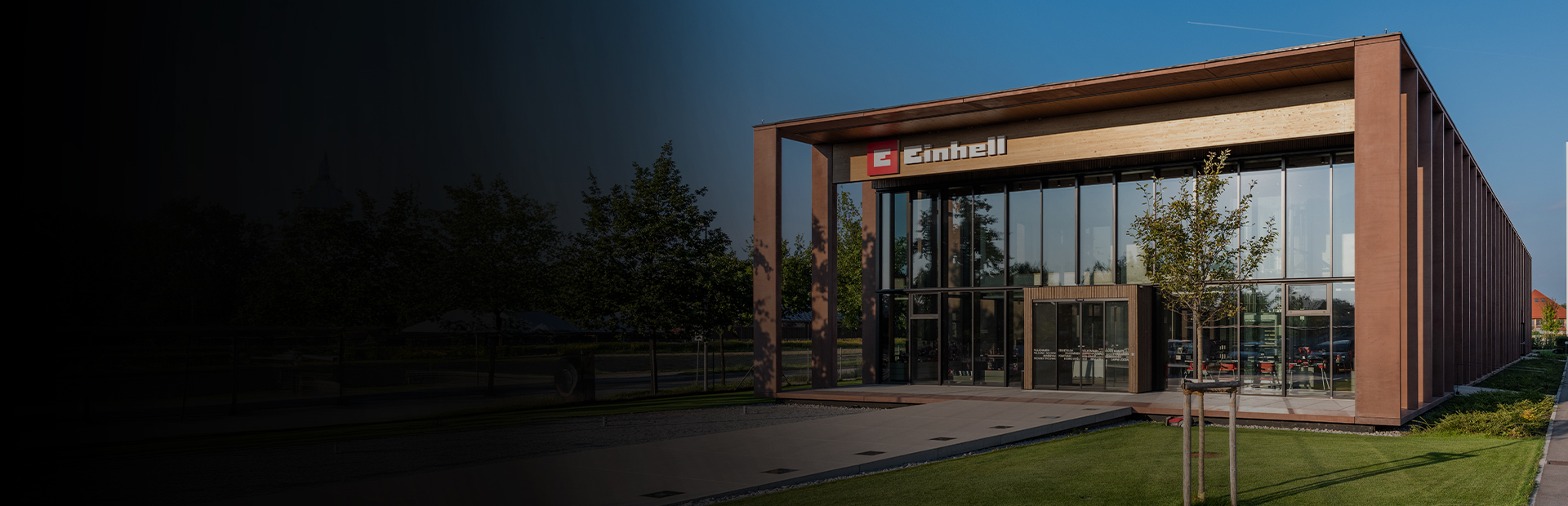 Modern Einhell building with glass facade and large logo. In front, trees, lawn, and a clear sky.