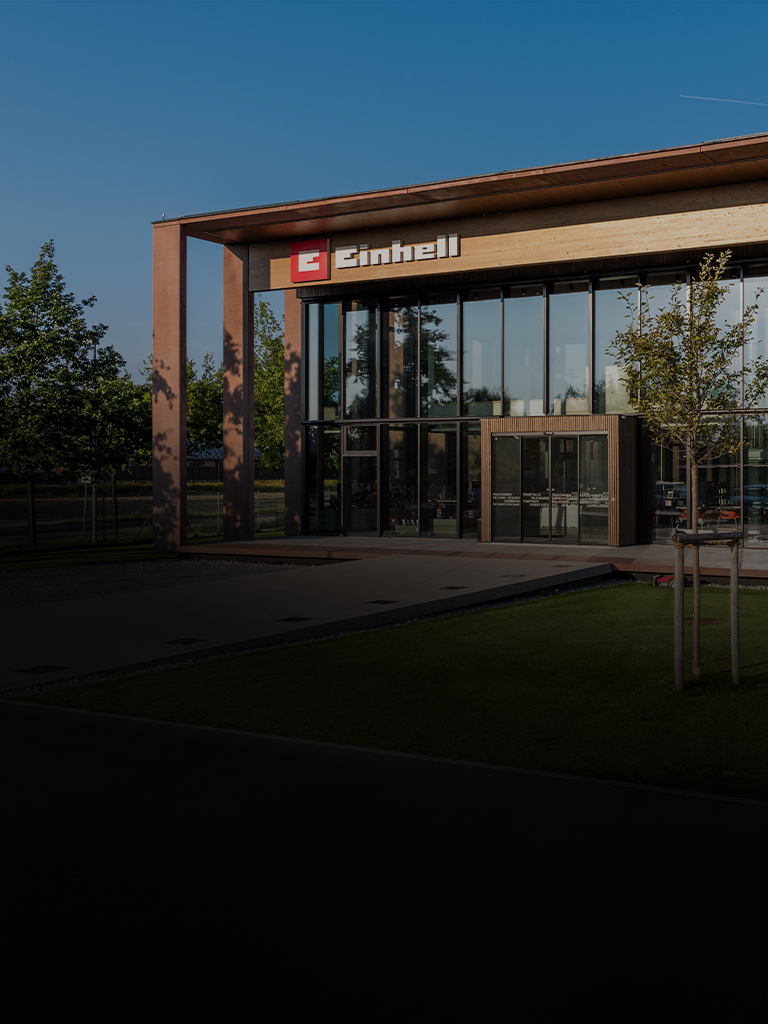 Modern Einhell building with glass facade and large logo. In front, trees, lawn, and a clear sky.