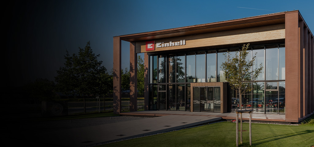 Modern Einhell building with glass facade and large logo. In front, trees, lawn, and a clear sky.