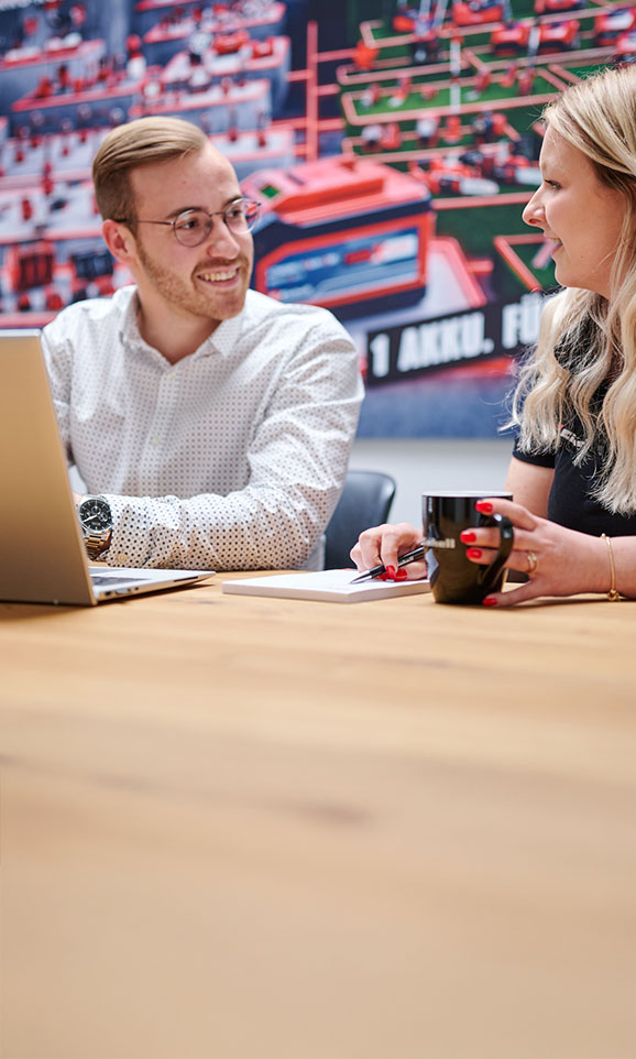 Two young colleagues are sitting at a table in an office, having a friendly conversation and working together on a laptop. In the background, a large Einhell poster featuring battery-powered products is visible.