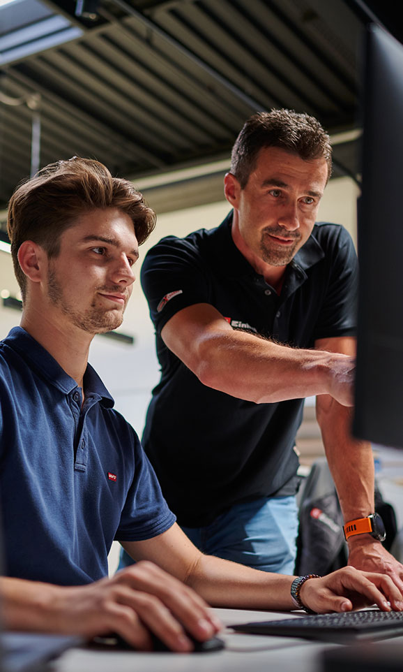A young man works at a computer while an older colleague points at the screen – teamwork and mentoring in a modern office.