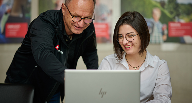 An older man points something out to a young woman on a laptop. Both are smiling.