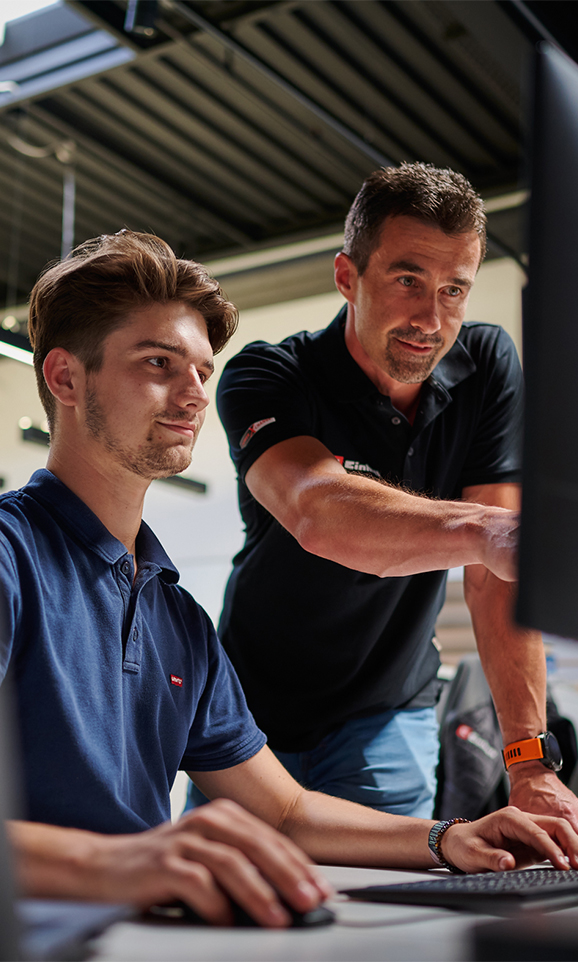 A young man and an older man working at a computer. The older man is pointing at the screen.