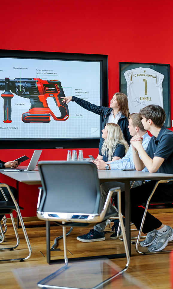 A woman is pointing at a screen with technical details of an Einhell rotary hammer. Four people are watching.