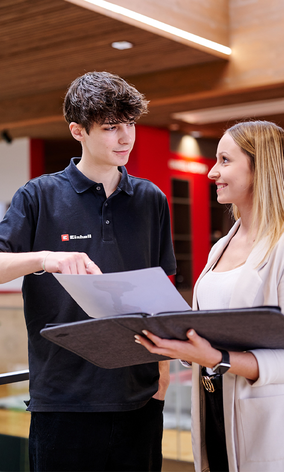 Two young Einhell employees in conversation. They are looking together at documents in an open folder.