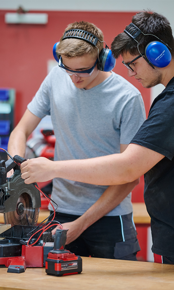 Two men wearing hearing protection work on a machine with an Einhell battery. They look focused.
